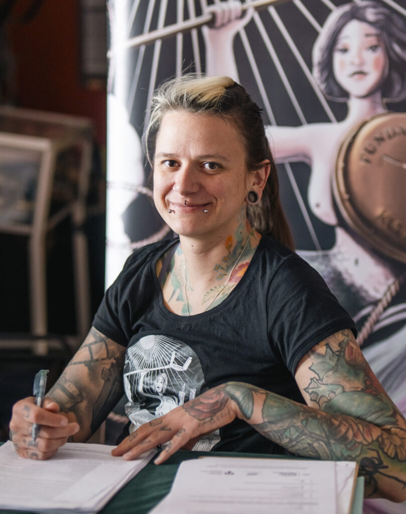 Zdjęcie uśmiechniętej Izabeli Sopalskiej Rybak siedzącej przy stole z dokumentami. Z nią widać rysunek syrenki warszawskiej na wózku z tarczą z napisem Fundacja Kulawa Warszawa