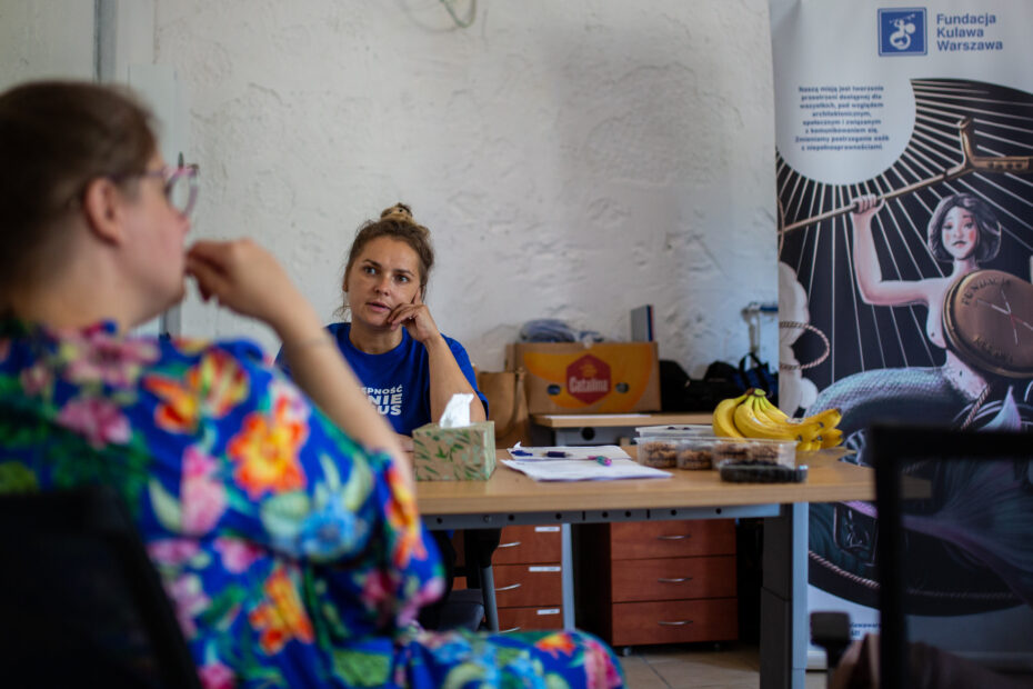 Anna Majdak rozmawia z uczestniczką spotkania. są w jasnym pomieszczeniu ze społem na którym stoją owoce. Obok stoi rollu fundacji kulawa warszawa
