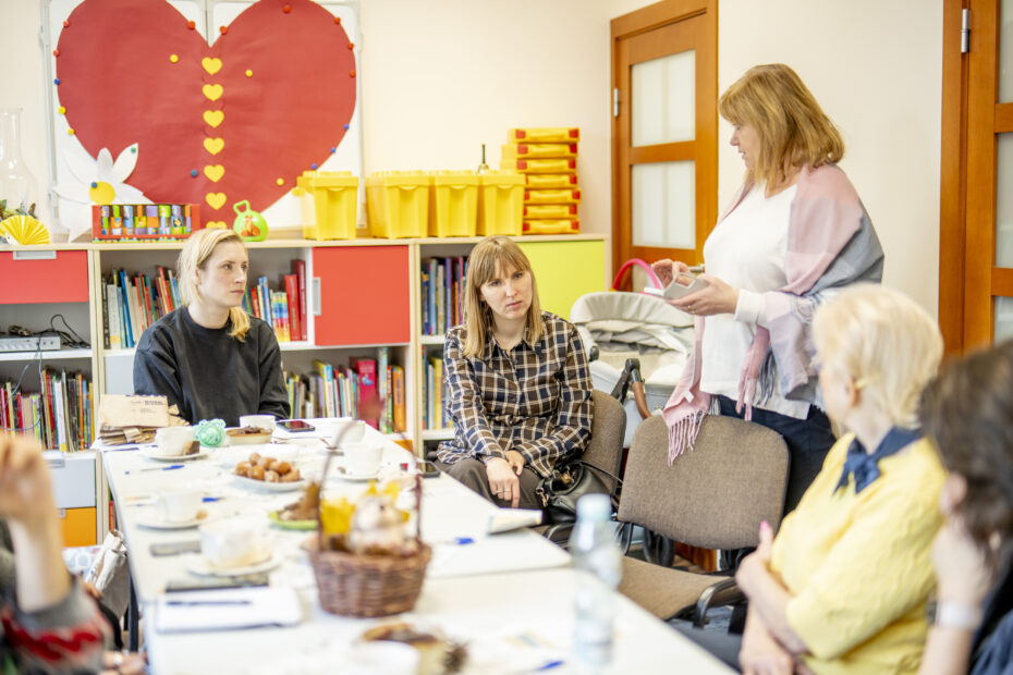 Przy stole z poczęstunkiem siedzi dużo osób. Na końcu stołu jest osoba niewidoma, a obok niej stoi osoba, z którą rozmawia. Pozostałe osoby siedzące przy stole słuchając rozmowy