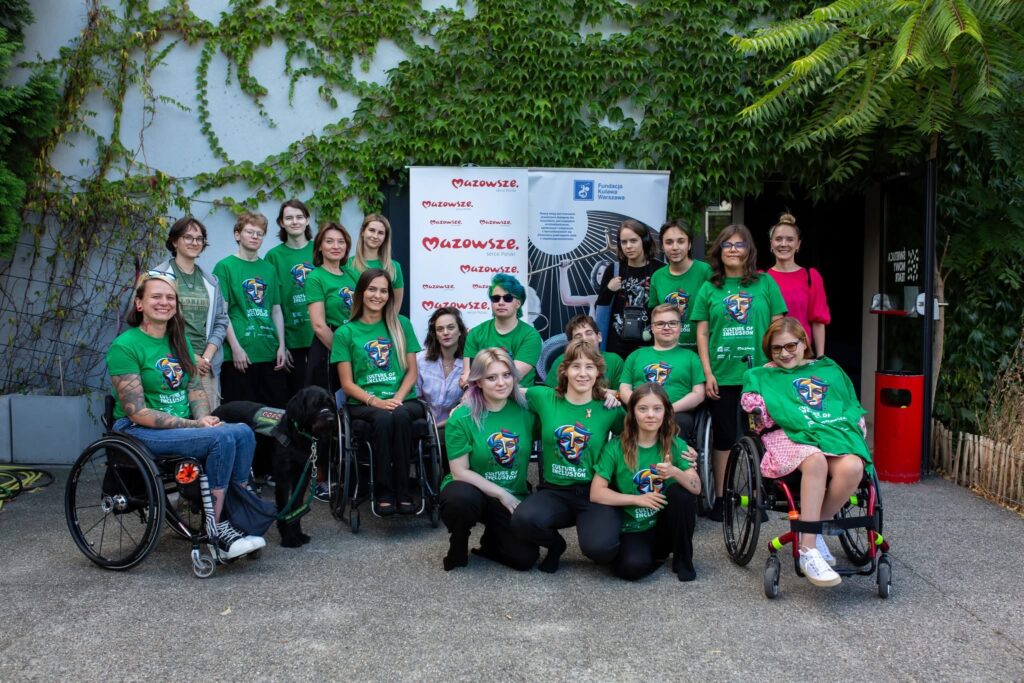 Osoby uczestniczące oraz organizatorki w zielonych koszulkach z logo projektu. Wszyscy stoją przed budynkiem świetlicy Nowego Teatru oraz przed rollupami fundacji Kulawa Warszawa i Mazowsza